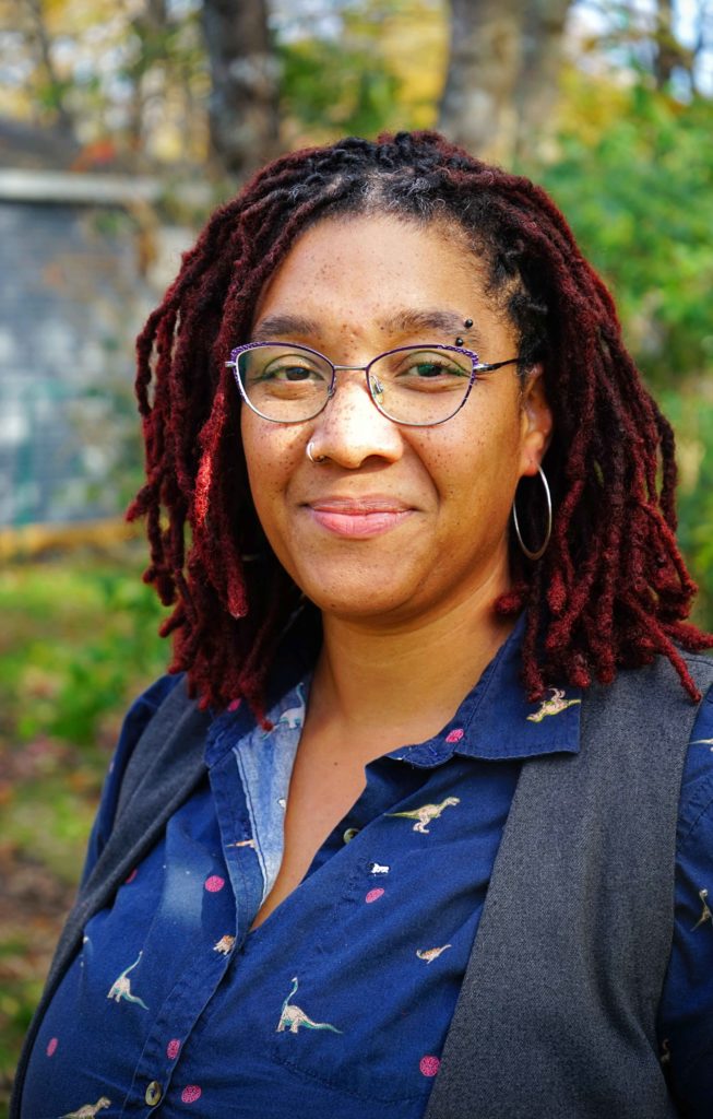 Ashley Miller is a Black femme person in their mid-thirties with shoulder length red locks, freckles and wearing glasses. She stands in front of a background with blurred treeds and greenery in her bark yard.